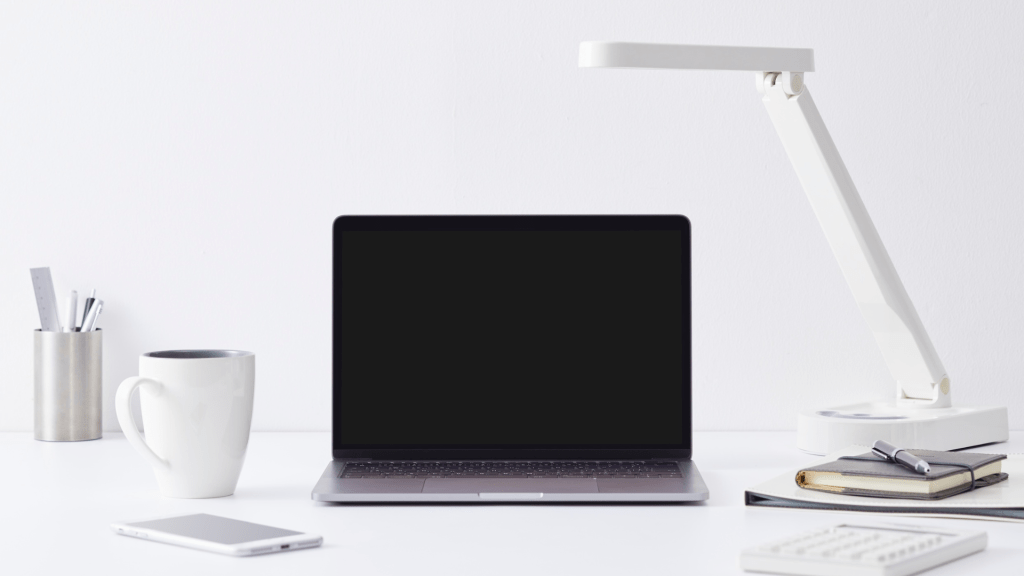 Photo of a white desk with a laptop, lamp, mug, phone, notebooks, and pen pot